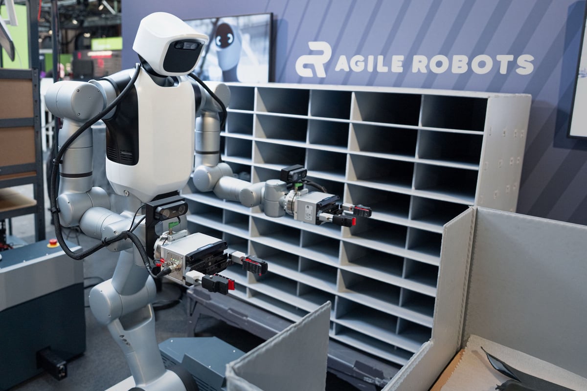 Robô humanoide da Agile Robots com dois braços junto a uma estante de compartimentos num stand.